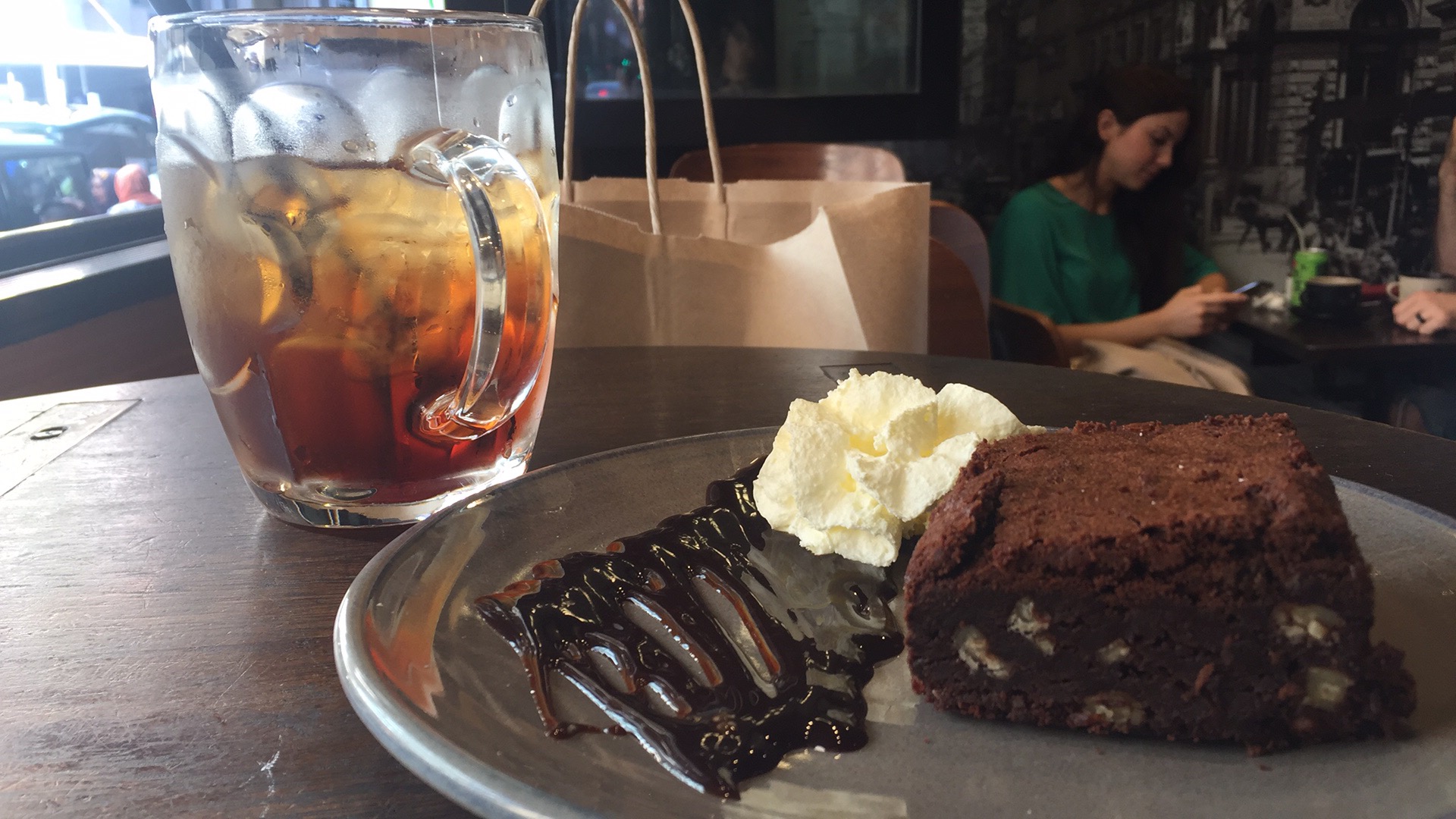 A plate with a brownie, and a glass mug of iced tea; a woman looks at her phone in the background