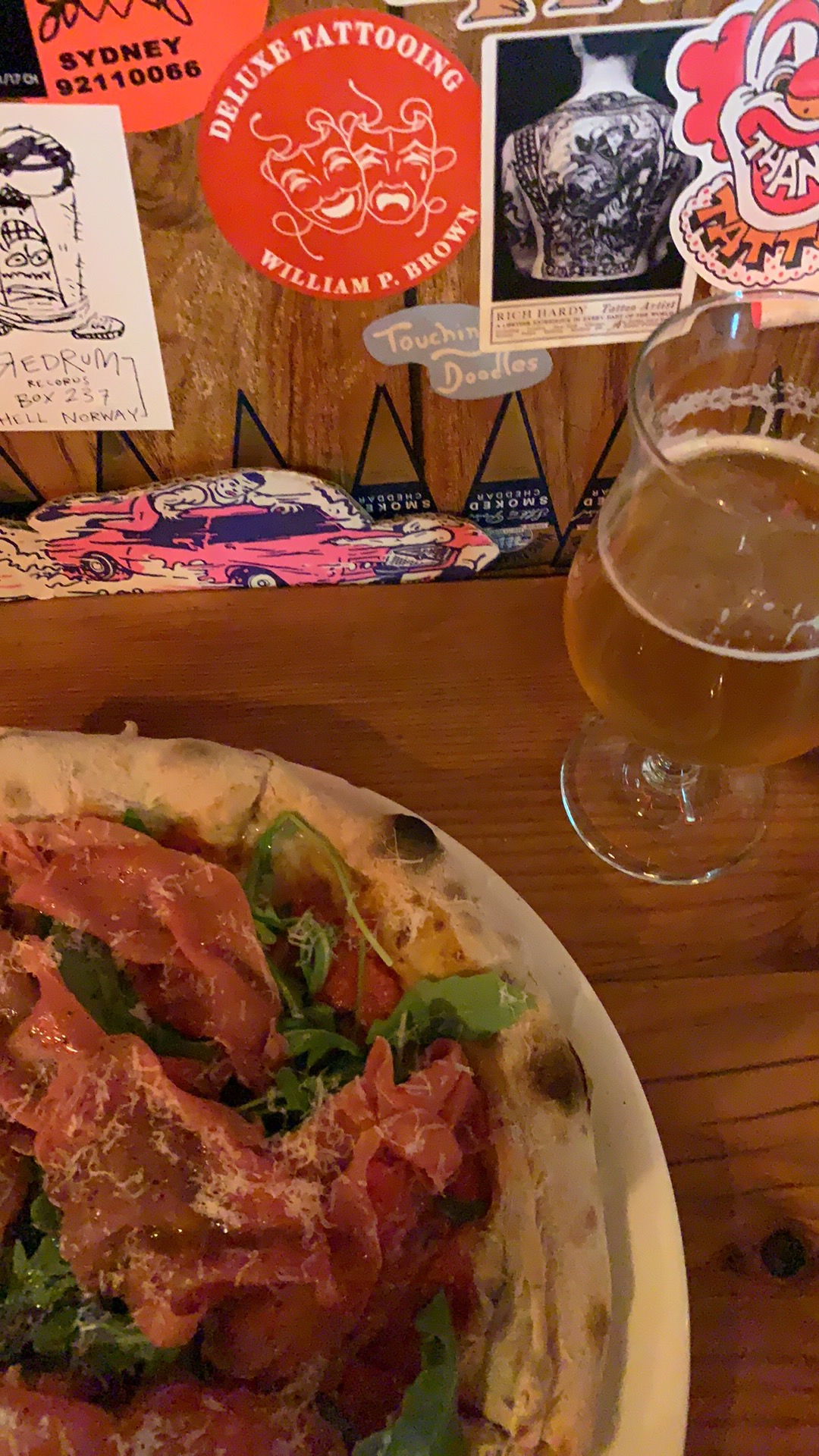 Pizza on a restaurant table, with a glass of beer; various stickers on the wall next to the table